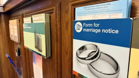 A number of plastic boxes attached a dark wooden panelling. Closest is blue, containing forms for marriage notices.