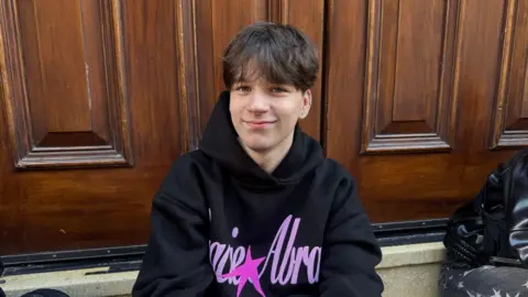 A young man sits in front of a large wooden door. He is smiling, wearing a black "Gracie Abrams" hoodie and he has brown hair with a fringe covering his forehead.