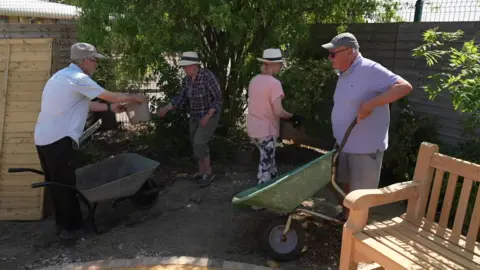 Three men and one woman dressed are moving plants with wheelbarrows 