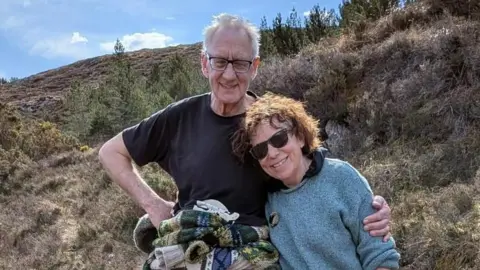 Hughie Stirling and Caroline Williams are pictured outdoors on a heather-covered hill. Hughie, who has his arm around Caroline, has grey hair, glasses and is wearing a black T-shirt. Caroline is wearing sunglasses and light blue jumper. They are smiling as they look into the camera.