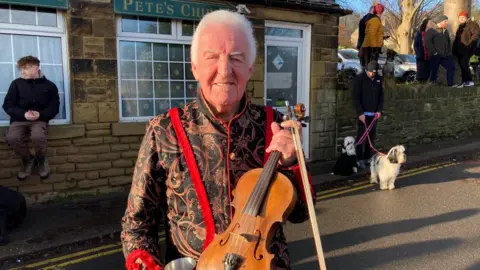 Ray Ellison stands outside the Old Harrow Inn, a traditional-looking stone-built pub, in Sheffield. He is holding a fiddle and bow and wearing a traditional sword-dancing jacket, which is black with an elaborate paisley design in shades of red and white.