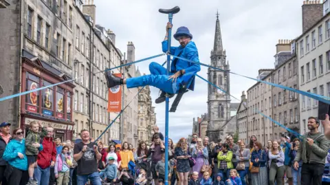 A performer outdoors on the street at the Edinburgh festival - he is dressed in a blue suit with a blue hat, and is at the top of a pole, with ropes being held by laughing people. The watching crowd are mostly all laughing as well.