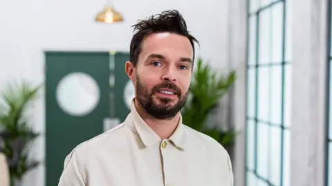 BBC/Optomen Television/Ashleigh Brown Oliver Jeffers. He has dark hair and beard, wearing a cream shirt.