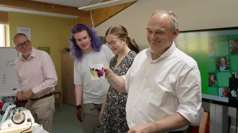Four people stand around a table during a workshop or demonstration. One person holds up a mug decorated with colourful sheep while others look on, with equipment and a presentation screen visible in the room.
