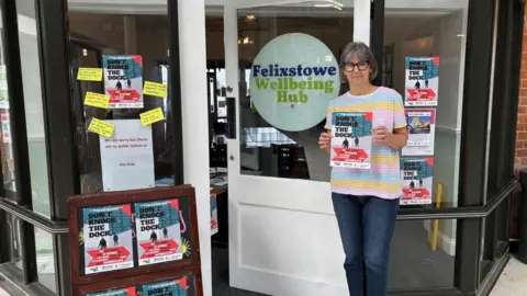 Karen Goddard Karen stands in front of the Felixstowe Wellbeing Hub. The windows are covered in posters for her play Don't Knock the Dockers. Karen is wearing a blue, yellow and pink striped T-shirt and jeans. She has glasses and bobbed dark grey hair