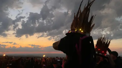 A picture of an animal with a hat on its head. The sunrise can be seen in the background alongside a large crowd.