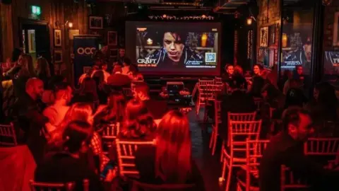 Siân Parry A group of people sitting on chairs with the back to the camera. They are facing a large screen. The lighting is red. On the screen there are various words, the only one which can made out being 'CRAFT'. 