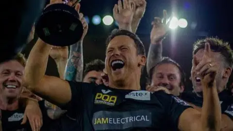 Joe Swash holding a trophy aloft with other men surrounding him