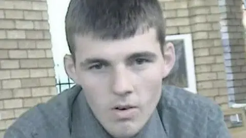 A photo of Anthony Atkins wearing a grey shirt, behind him is a brown bricked building 