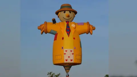 Longleat Large scarecrow-shaped hot air balloon in the sky. It has a carrot for a nose, and short blonde hair poking out from beneath a brown straw hat with a green band. It is wearing an orange coat with red, green and purple patches sewn on. It has a red tie with blue circles and a yellow shirt. The balloon design cuts off just below the waist but its lower garment is white with red hearts. A crow is sitting on its shoulder.