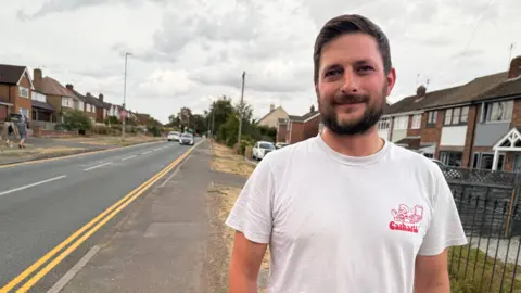 A man with dark hair and a beard wearing a white t-shirt. He is standing on a pavement and next to him you can see freshly painted double yellow lines on the road. There are houses on either side of the road.