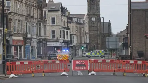 Road closed sign next to a police cordon. Police vehicles are parked next to the area. 