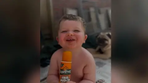 Claire Barlow Picture of Osian smiling looking at the camera holding an orange ice lolly. He has short brown hair and is not wearing a top. He is sat in his garden. Behind him is a pug who is sat down.