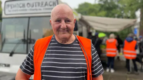 Ian Walters stands in a high-vis vest in front of a bus. There are people gathered to get food from the bus.