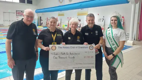Grantham Rotary Club A group of people stood smiling and holding a large cheque in front of a laned swimming pool. Two women and two men are wearing black polo shirts with a yellow circular logo on it. On the right is a woman with turquoise hair wearing a tiara and a ribbon saying 'Ms Earth UK' on it. The cheque says The Rotary Club of Grantham, Lincs & Notts Air Ambulance, £1,000.