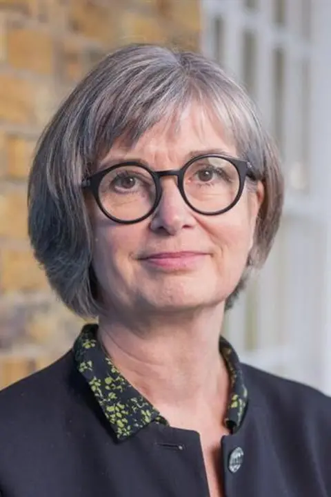 RCN Patricia Marquis is looking directly at the camera. She has a short grey bob and is wearing rounded, dark-rimmed glasses and a dark jacket. 