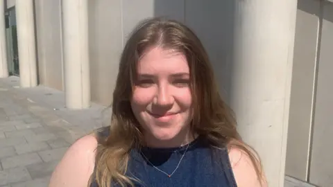A girl with long light brown hair, wearing a blue sleeveless top and silver neckless and smiling while looking at the camera.