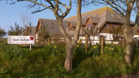Sark school, with a sign saying "welcome to Sark school", and trees and flowers infront of it.