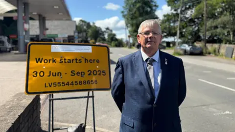 Patrick Allen, a man wearing a navy blue suit, is stood next to a yellow road sign with black writing with details of road works. There is a petrol station forecourt behind him.