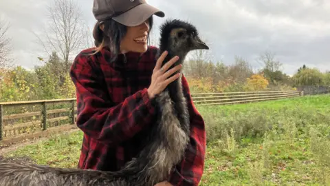 Rhi Evans cuddles an emu in a field. Ms Evans is wearing a black baseball cap and a red plaid shirt. 