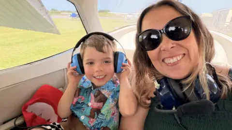 An image of Nicki Hale and her son sat in a small plane. They are both smiling at the camera. Nicki has a pair of headphones around her neck, and her son has them on his head. He looks about four years old and is wearing a brightly coloured shirt. 
