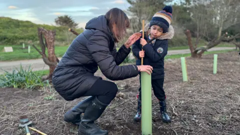 Woman in her thirties, she has a long brunette bob and is crouched down by her son as they plant a tree together. 