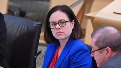 Getty Images Kate Forbes sits in the chamber at Holyrood. She has shoulder length dark brown hair and wears a red shirt, blue blazer and black glasses.