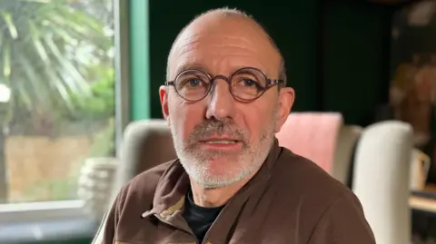 Simon Bessell is wearing round black glasses and a brown sweatshirt. He has a grey beard. 