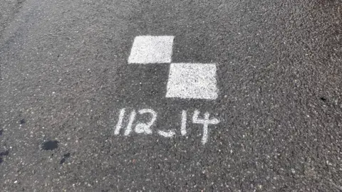 Two white squares painted on a road. Underneath is the number "112_14".