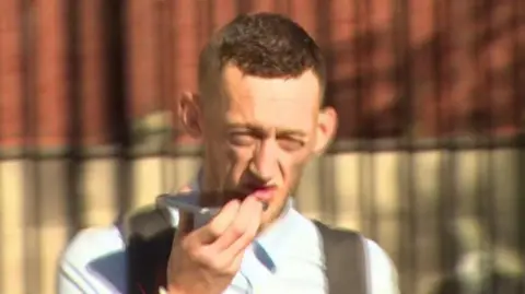 BBC Craig Rowland - a still of a man with short brown hair wearing a light blue collared shirt and backpack as he holds a phone up to his mouth. He is squinting his eyes as he looks at the camera. He is walking in front of a tall, black gate and a red brick building.