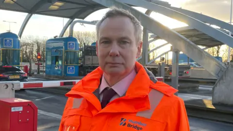 A man with short fair hair looks into the camera as he stands in front of toll booths at the entrance to a bridge. He is wearing a hi-vis orange jacked over a suit, lilac shirt and dark tie. Behind him is a red and white barrier blocking a road. A blue toll booth and red lights can also be seen. A truck is waiting at the toll booth. The sun is low in the sky.
