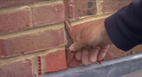 An exterior brick wall of a house has a loose mastic joint. A man's hand pulls the seal, which sits between two lines of bricks, away from the wall