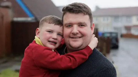 PA Media A small boy puts his hands round a man with short brown hair. The boy has short brown hair and a red jumper and a yellow polo. The man has a black sweatshirt on. Behind them is an out-of-focus street. The sky is grey. 