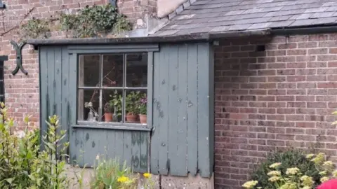 National Trust Wooden clad small porch attached to house with window in its centre - plants and flowers can been seen at its entrance.