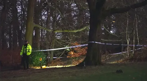 A police officer in a high viz jacket and police tape around a wooded area