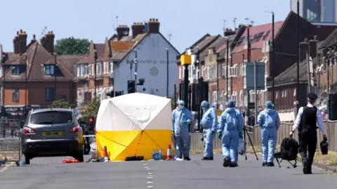 A forensic tent stands in the middle of a residential road, surrounded by several people in protective suits and police cordons. Parked vehicles line the street, while rows of brick houses and traffic lights are visible in the background on a clear day.