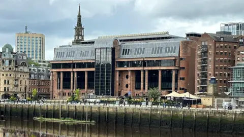 Newcastle Crown Court reflected in the River Tyne running in front of it. It is an imposing building made from smooth red stone with massive black windows and tall columns along its frontage.