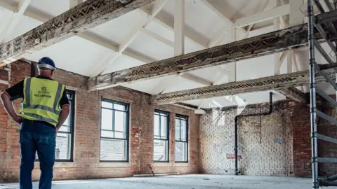 Lucille Moore A man in a yellow high-vis vest and a hard hat and dark trousers stands with his back to the camera looking at a room with beams and exposed brick work. There is scaffolding to the right of the picture and the floor is dusty
