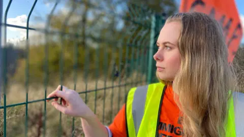 Emma Pearson, wearing an orange jumper and yellow hi-vis jacket, looks through a wire fence at the detention centre site. She has long blonde hair and a nose ring.