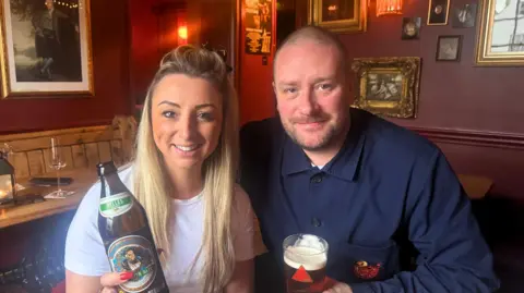 BBC/ Emily Johnson Two people sit together in a warmly lit pub with deep red walls and framed artwork. The woman on the left holds a bottle of beer, while the man on the right has a pint glass of amber ale. They’re seated at a wooden table with red cushioned stools, surrounded by string lights and other bottles and glasses.