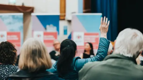 A woman wearing a blue coat raising her hand at a meeting, she faces ministers that have been blurred by the camera. She sits in between other people who are looking in the same direction.