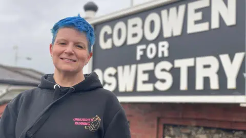 A blue-haired woman, wearing a black hoodie, standing in front of a black and white railway sign, which reads, "Gobowen for Oswestry".