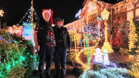 BBC A couple dressed in dark clothing stand outside a house with Christmas lights surrounding them. The man on the left is wearing a red Santa hat and a T-shirt with green, red and white writing on it. The woman on the right is stood wearing a black coat with a green elf hat. There are green lights on a hedge to the side of them, light up candy canes lining the path and a golden street lamp among the hundreds of lights around them.