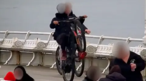 A bike rider does a wheelie along a pier with seats behind them and a number of  people in view.
