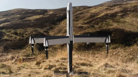 A four-winged drone plane, sitting at the bottom of a grassy hill. It is white and black and standing upright