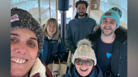Five people (three women and two men) all smiling at the camera taken as a selfie. Four of them are smiling with open mouths, and they are all wearing winter clothing with three of them wearing hats. Benches can be seen on both sides behind them, and outside there is deep snow and ice all around. 