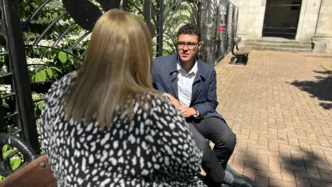BBC reporter Josh Sandiford speaks to the anonymous mother of a Swinfen Hall prisoner. He is wearing a blue jacket and a white shirt. She has a black and white dress on. We can only see the back of her head