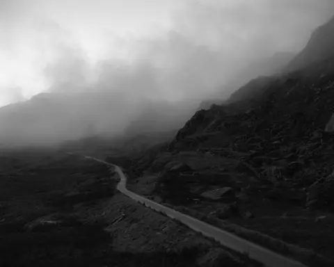 Kyle McDougall Kyle's black and white photograph of a mountain road with a craggy hillside on one side and grass sloping bank on the other. The sky above is misty white. 