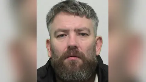 A police mugshot of James Murdoch, who has a beard with greying short hair and is wearing a dark jacket. 