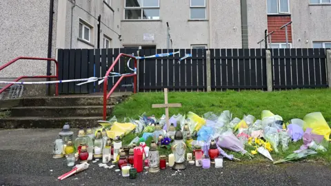 Pacemaker Candles, flowers, and a cross left on the ground outside a block of flats with the entrance covered by police tape